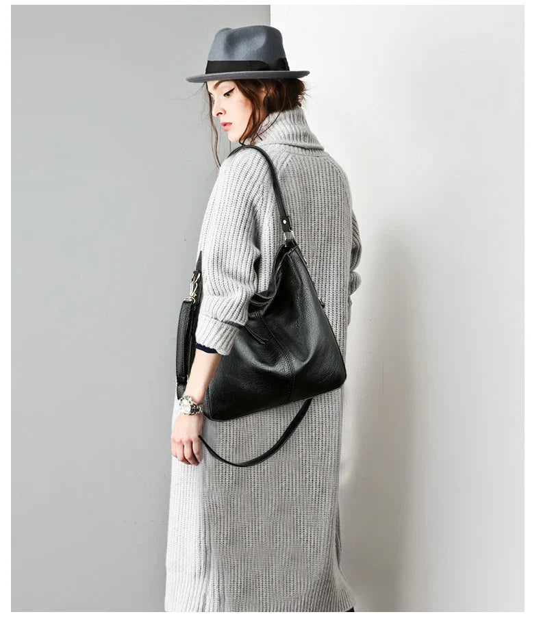 Casual black faux leather hobo bag worn over the shoulder, paired with a gray knitted dress and a gray hat.