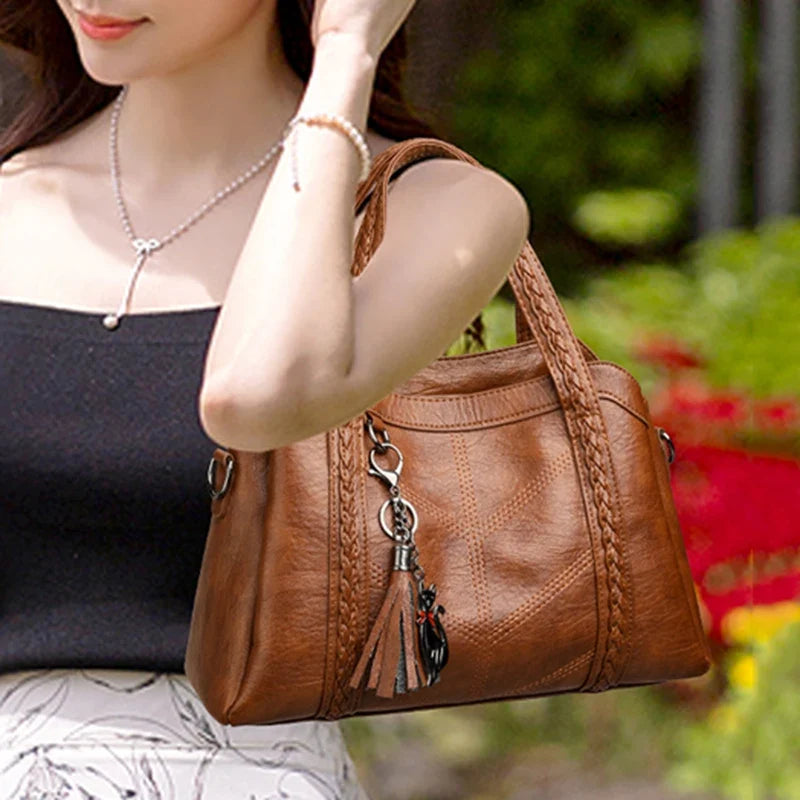 Brown leather shoulder bag with a braided handle and tassel charm, shown on a model wearing a black top.