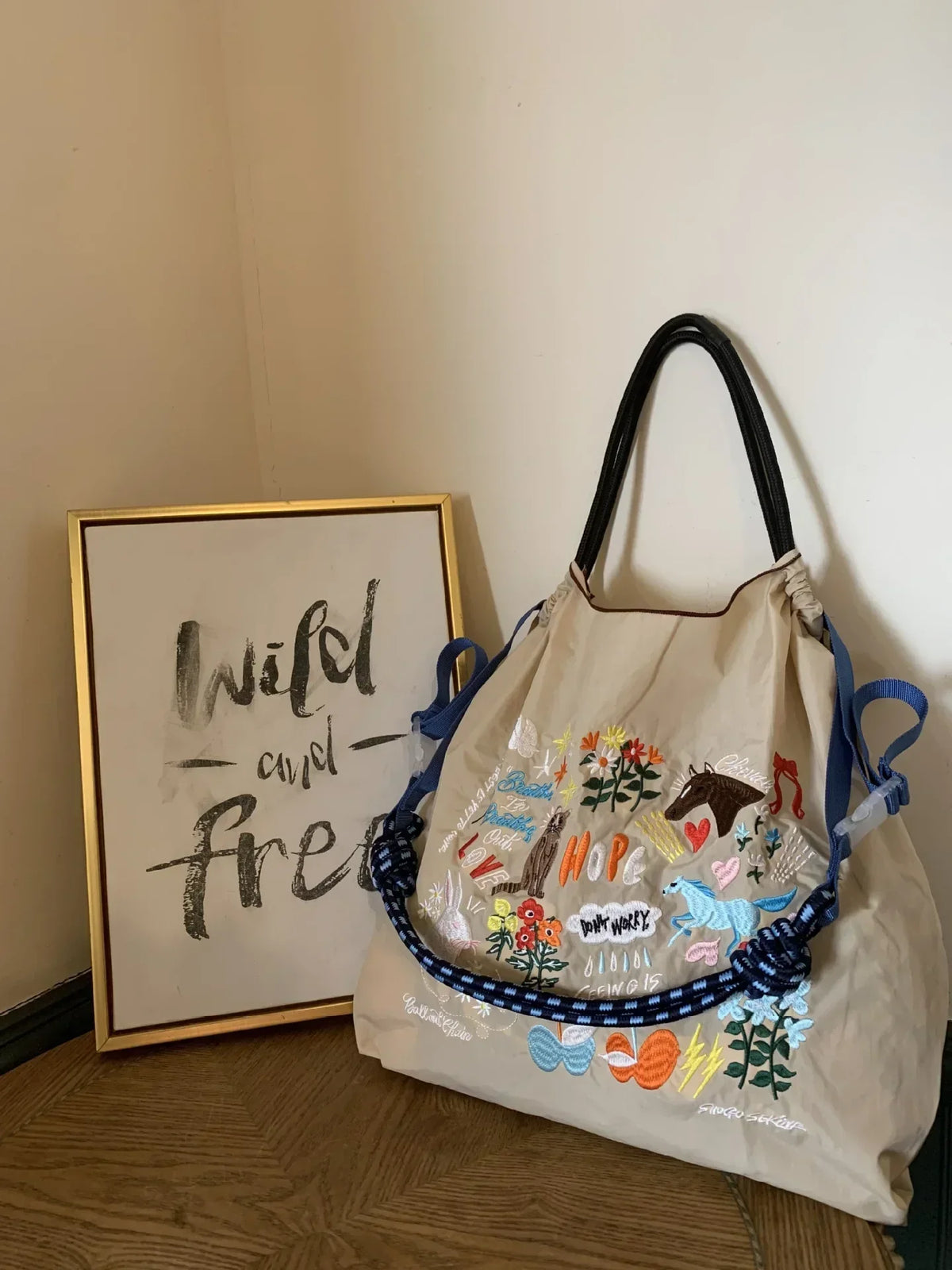 Large beige nylon tote bag featuring colorful embroidered designs, adjustable blue straps, and a casual style.