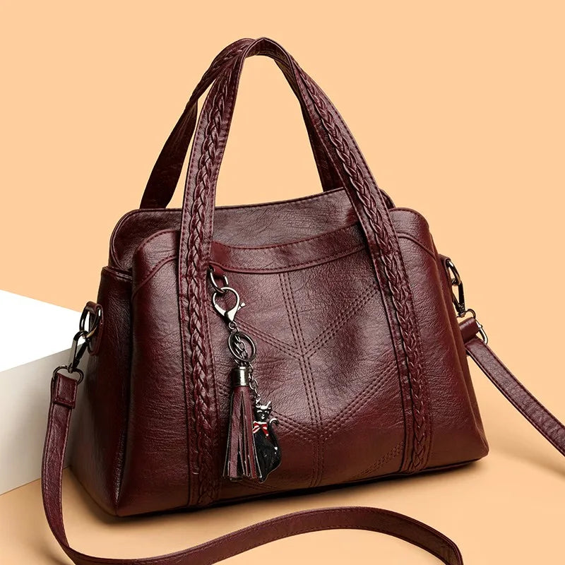Burgundy leather shoulder bag with braided handles, tassel charm, zip closure, and a casual tote shape.