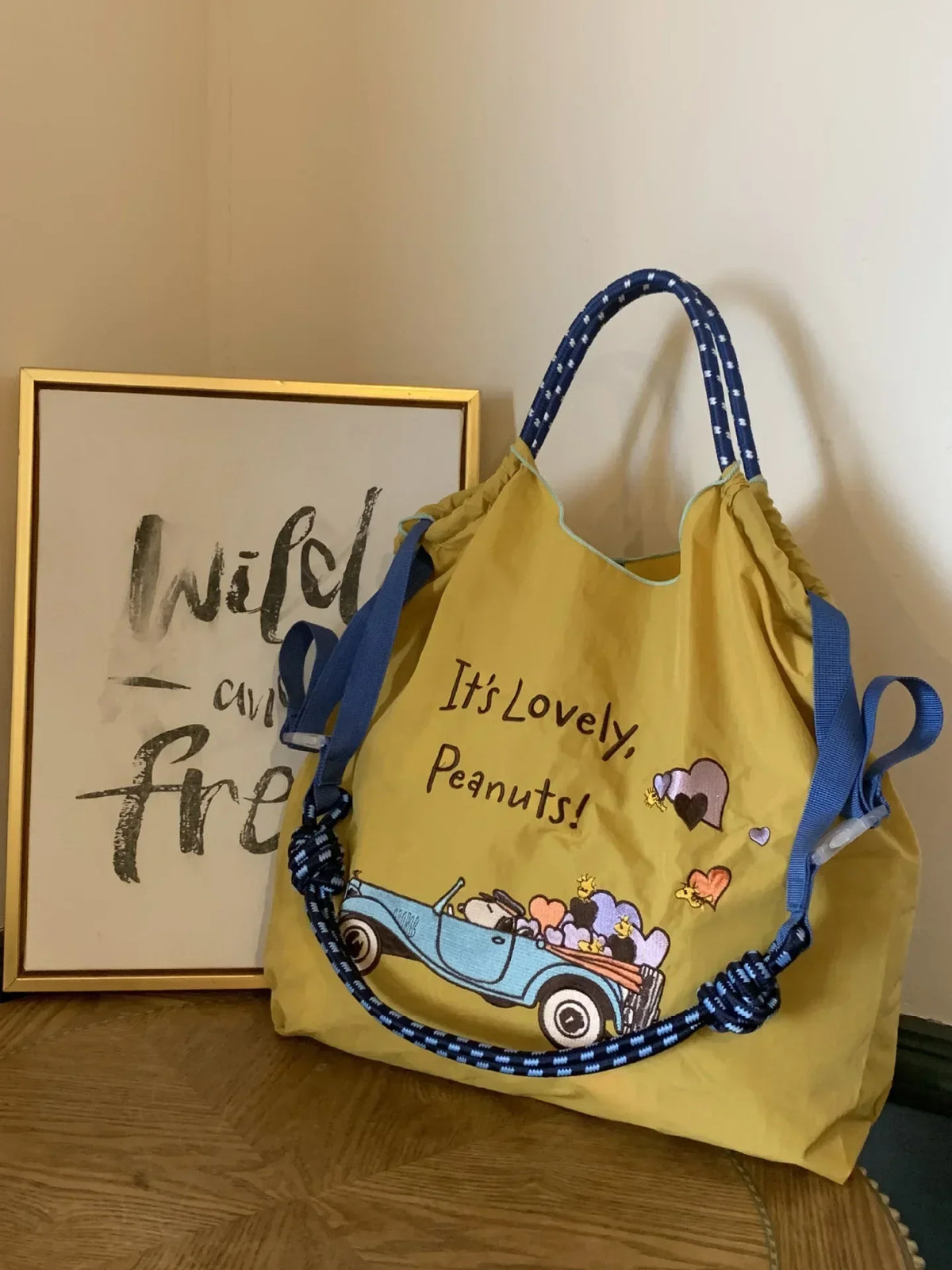 Yellow nylon tote bag featuring a playful design with the text "It's Lovely, Peanuts!" and a blue convertible graphic. Blue and white patterned handles.