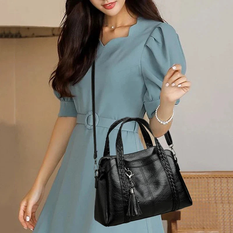 Black leather shoulder bag with braided handles and a tassel detail, worn crossbody. Model in a blue dress.