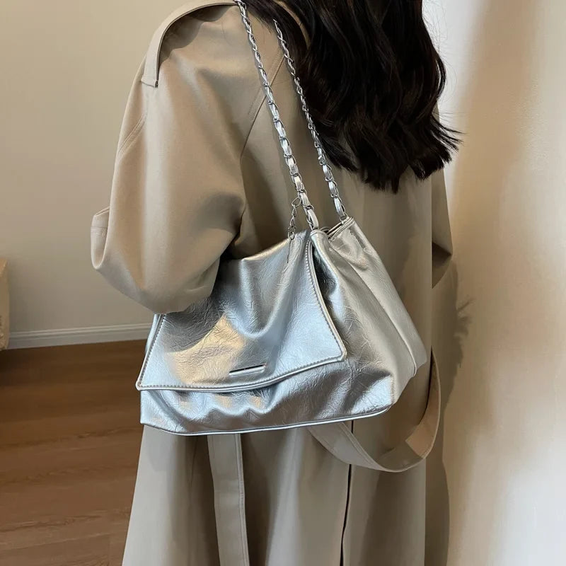 Silver shoulder bag with a shiny surface and chain strap, worn underarm, paired with a beige coat.