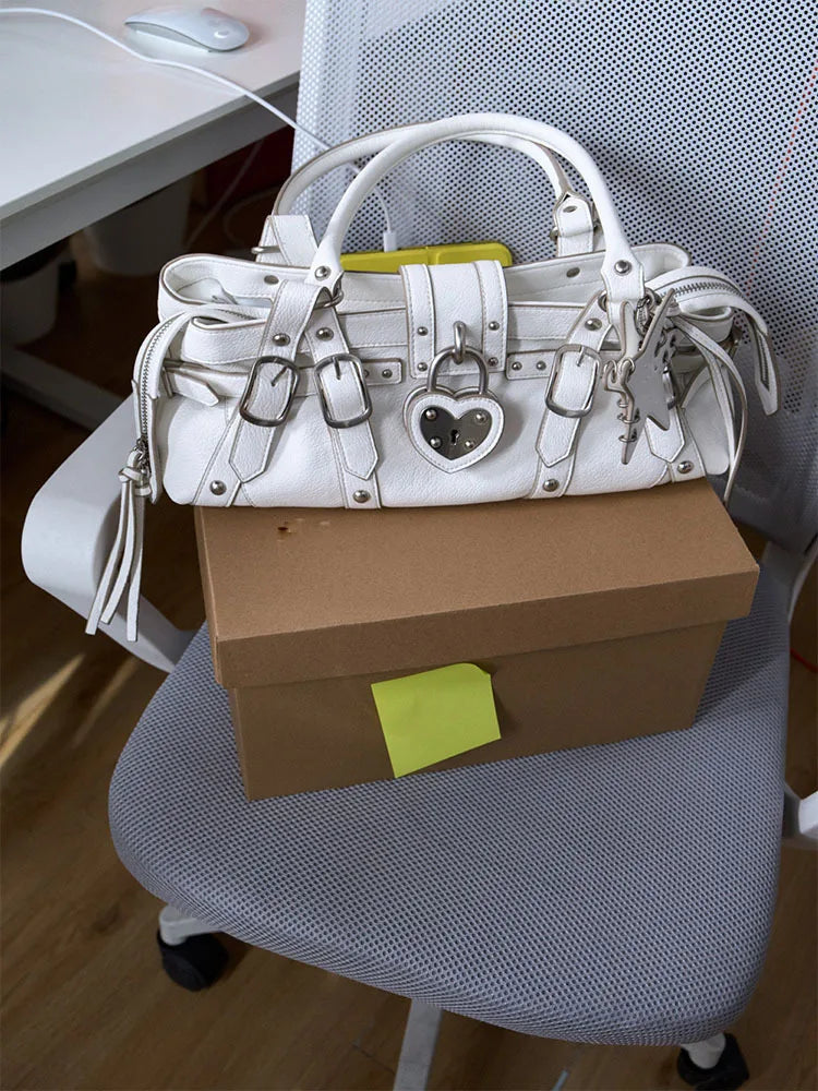White shoulder handbag with rivet details and a heart-shaped lock, resting on a brown box on a chair.