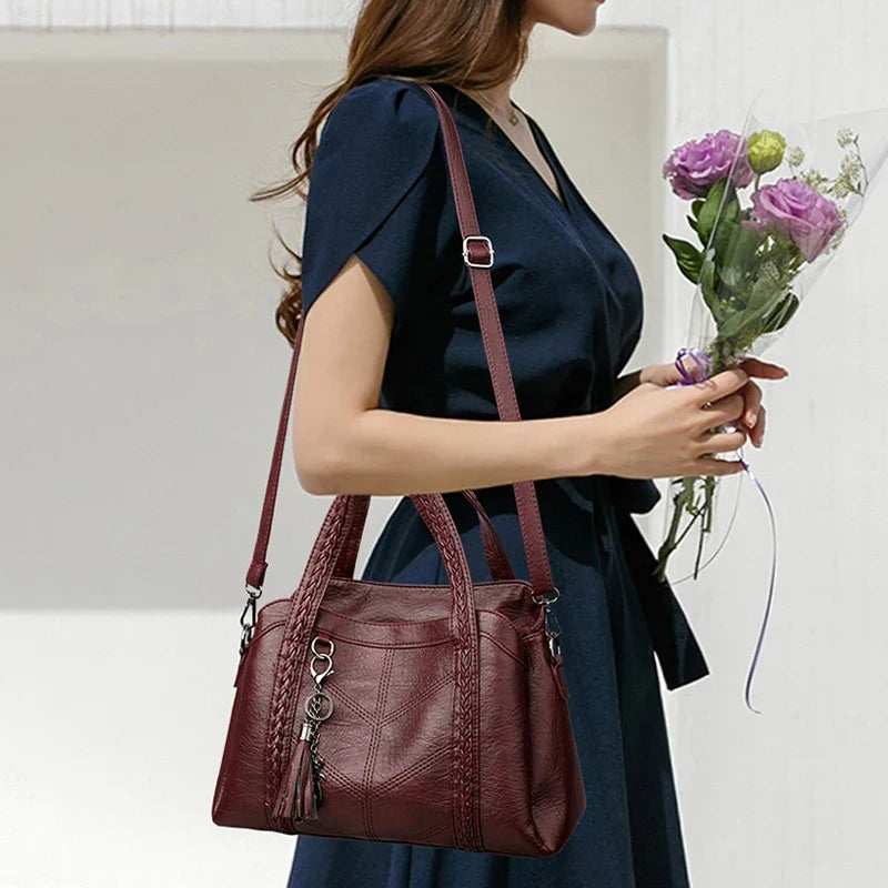 Burgundy leather shoulder bag with braided straps, worn crossbody style by a woman holding flowers, featuring a zip closure.