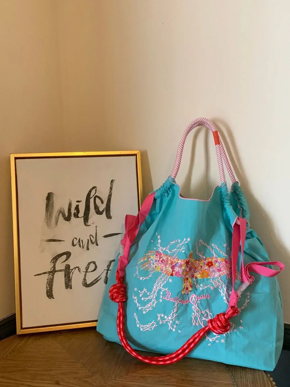 Light blue nylon tote bag with embroidered floral design, pink straps, large capacity, and casual style, placed beside a framed art print.