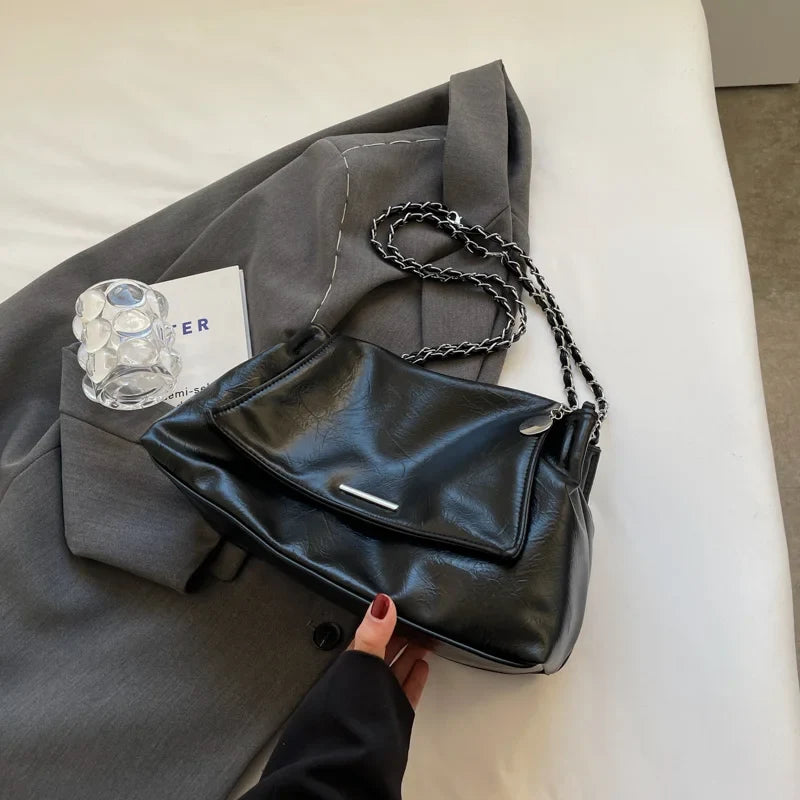 Black soft leather shoulder bag with a flap pocket and chain strap, resting on a gray blazer and styled with a notebook.