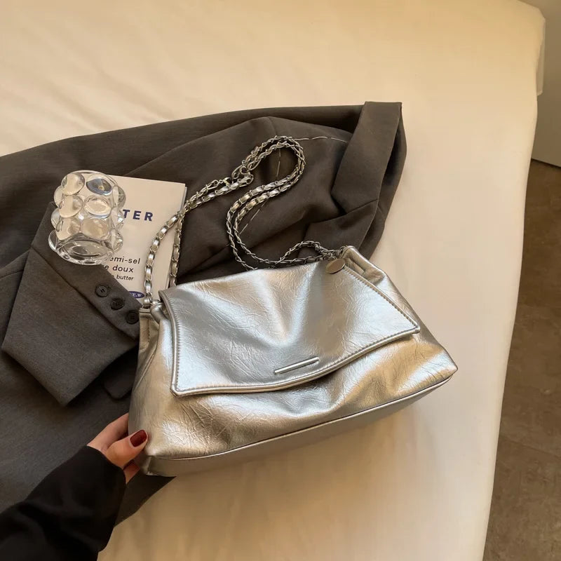 Silver soft leather shoulder bag with a flap pocket, featuring a chain strap, resting on a gray fabric.