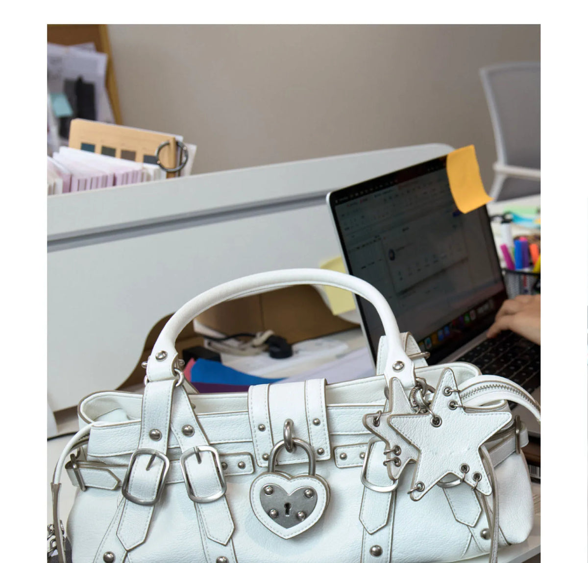 White crossbody handbag with rivet detailing, heart-shaped padlock, and star-shaped charms. Soft microfiber synthetic leather.