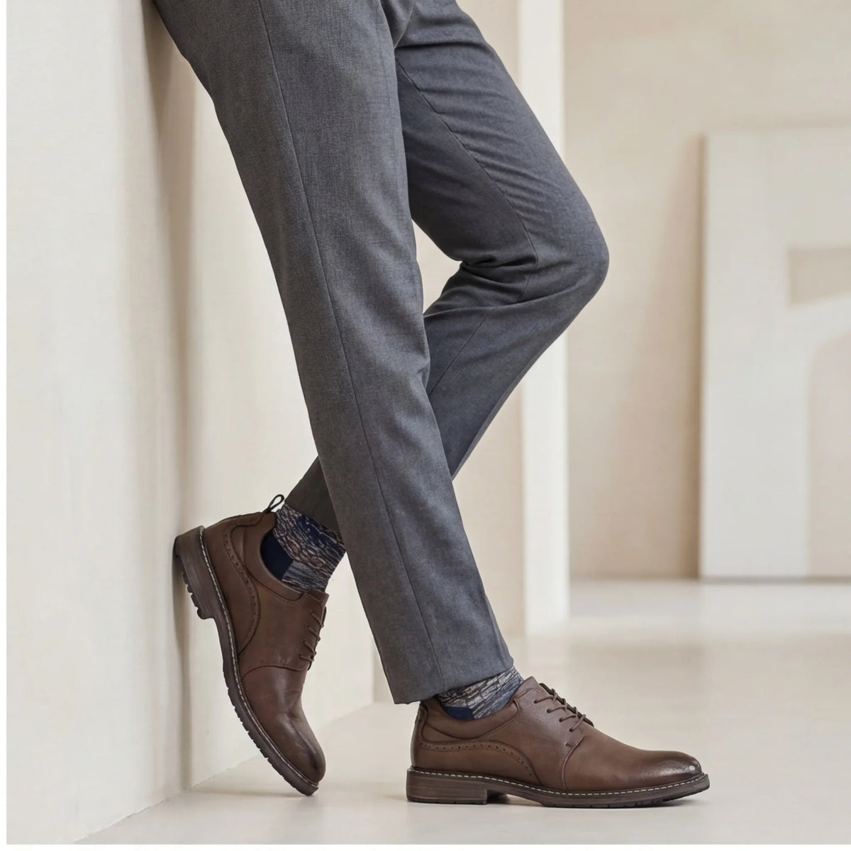 Brown lace-up derby shoes with a round toe, worn with gray trousers and patterned socks, showcasing a business casual style.