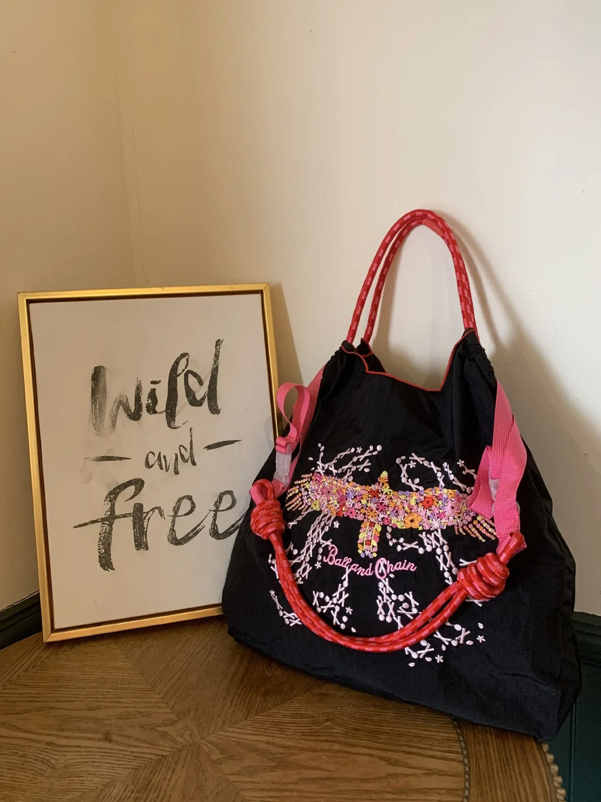 Large black nylon tote bag with pink embroidered design and matching rope handles, displayed next to a framed sign.