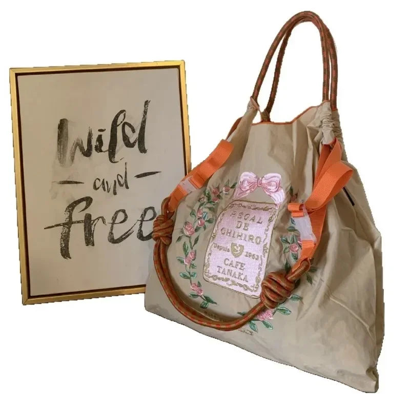 Lightweight beige tote bag with floral embroidery, orange straps, and a casual design, displayed next to a framed quote.