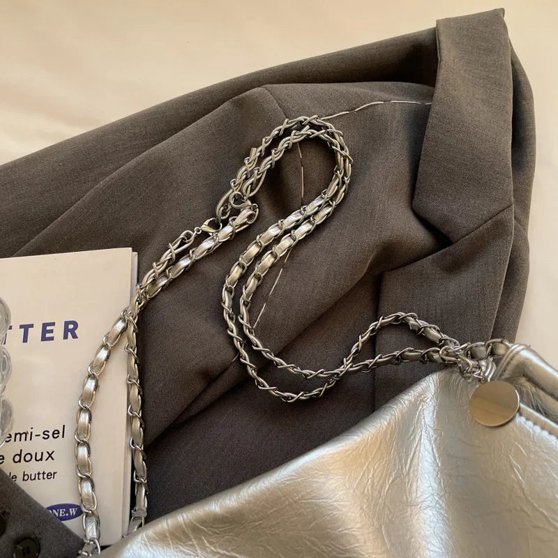 Silver shoulder handbag with a soft leather finish, chain strap, and spacious design resting on grey fabric.