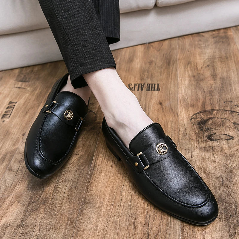 Men's black leather slip-on loafers with a round toe and a metal buckle detail, worn with tailored trousers on a wooden floor.