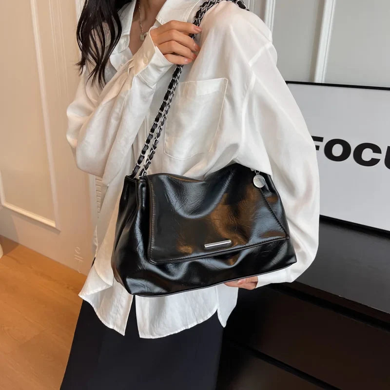 Black soft leather shoulder bag with a chain strap, held by a model in a white shirt, showcasing a casual tote style.