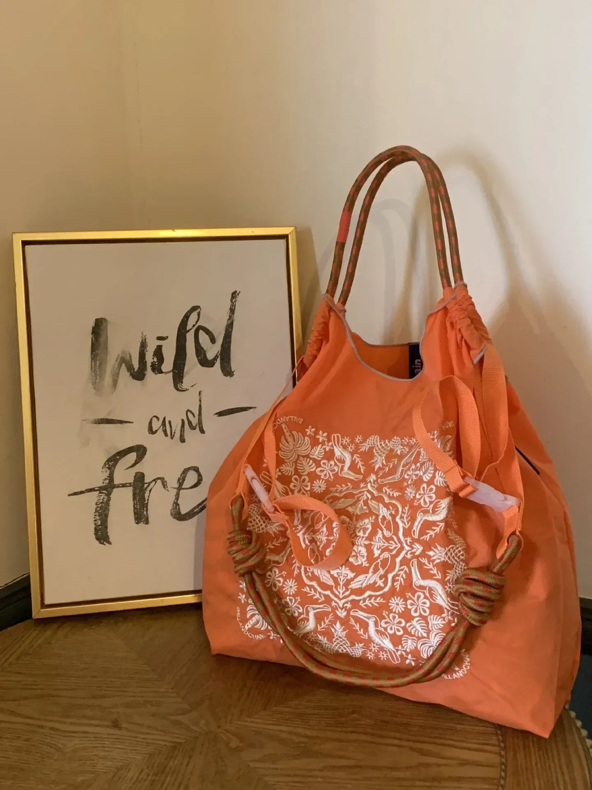 Large-capacity orange nylon tote bag with white floral embroidery and colorful twisted handle, placed near a framed quote.