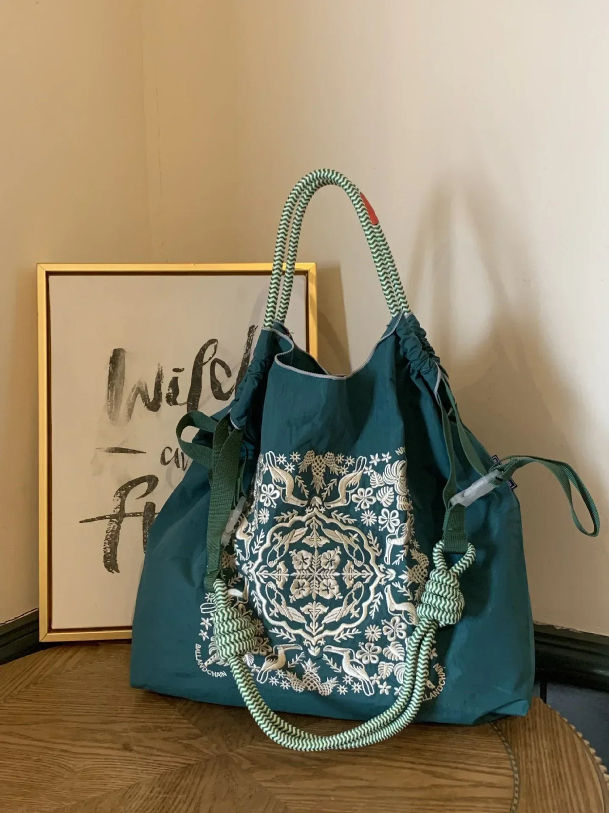 Large teal nylon tote bag with white floral embroidery, featuring braided green handles and a casual style.
