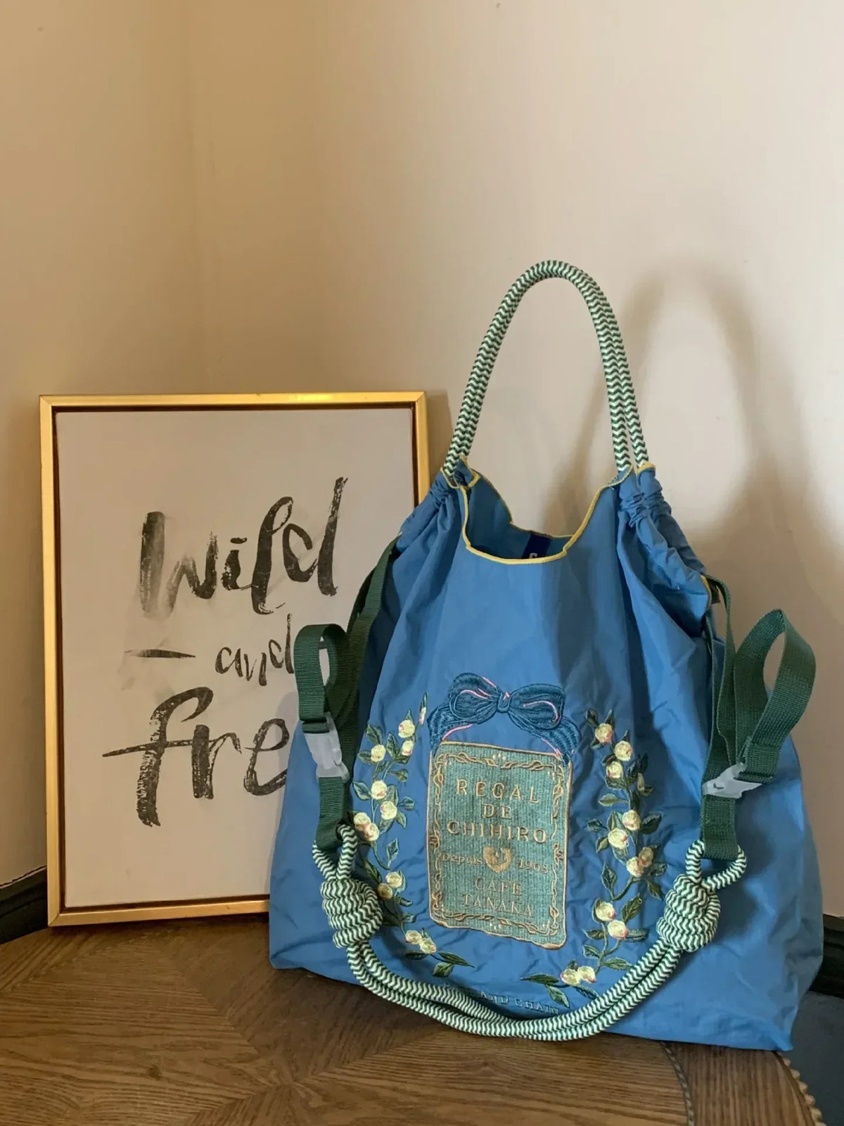 Large blue nylon tote bag with floral embroidery, green straps, and a drawstring closure, resting beside a framed quote.