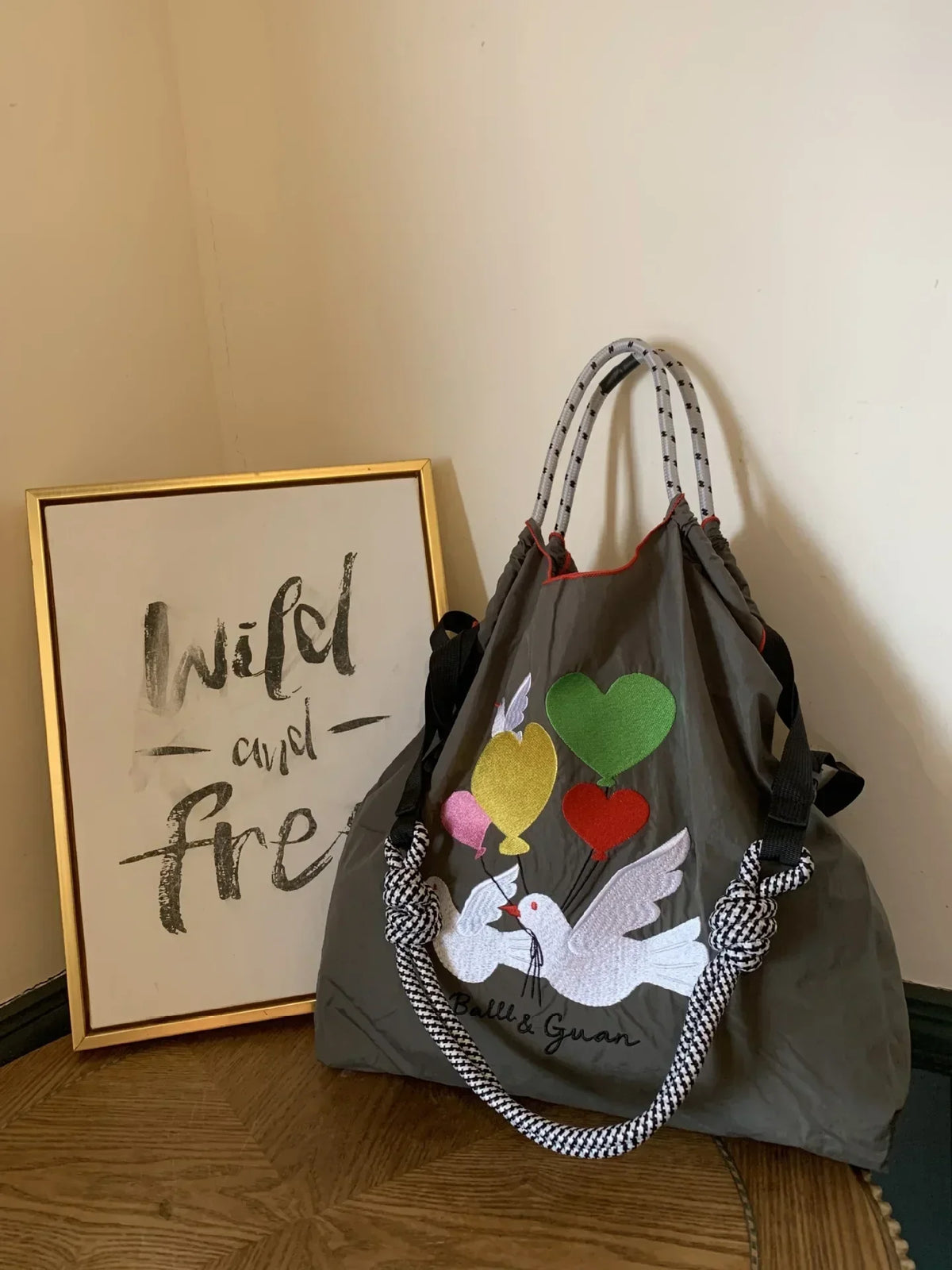 Large gray nylon tote bag features colorful embroidered doves and hearts, with a houndstooth-patterned handle.