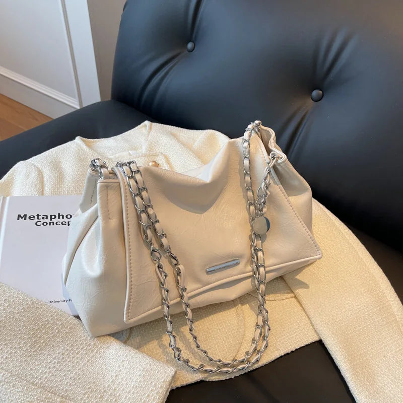 Soft beige shoulder bag with a silver chain strap, featuring a flap pocket and two handles, placed on a textured fabric surface.