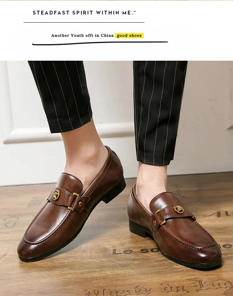 Brown leather slip-on loafers with a metal buckle, worn with pinstriped trousers. Rounded toe and casual business style.