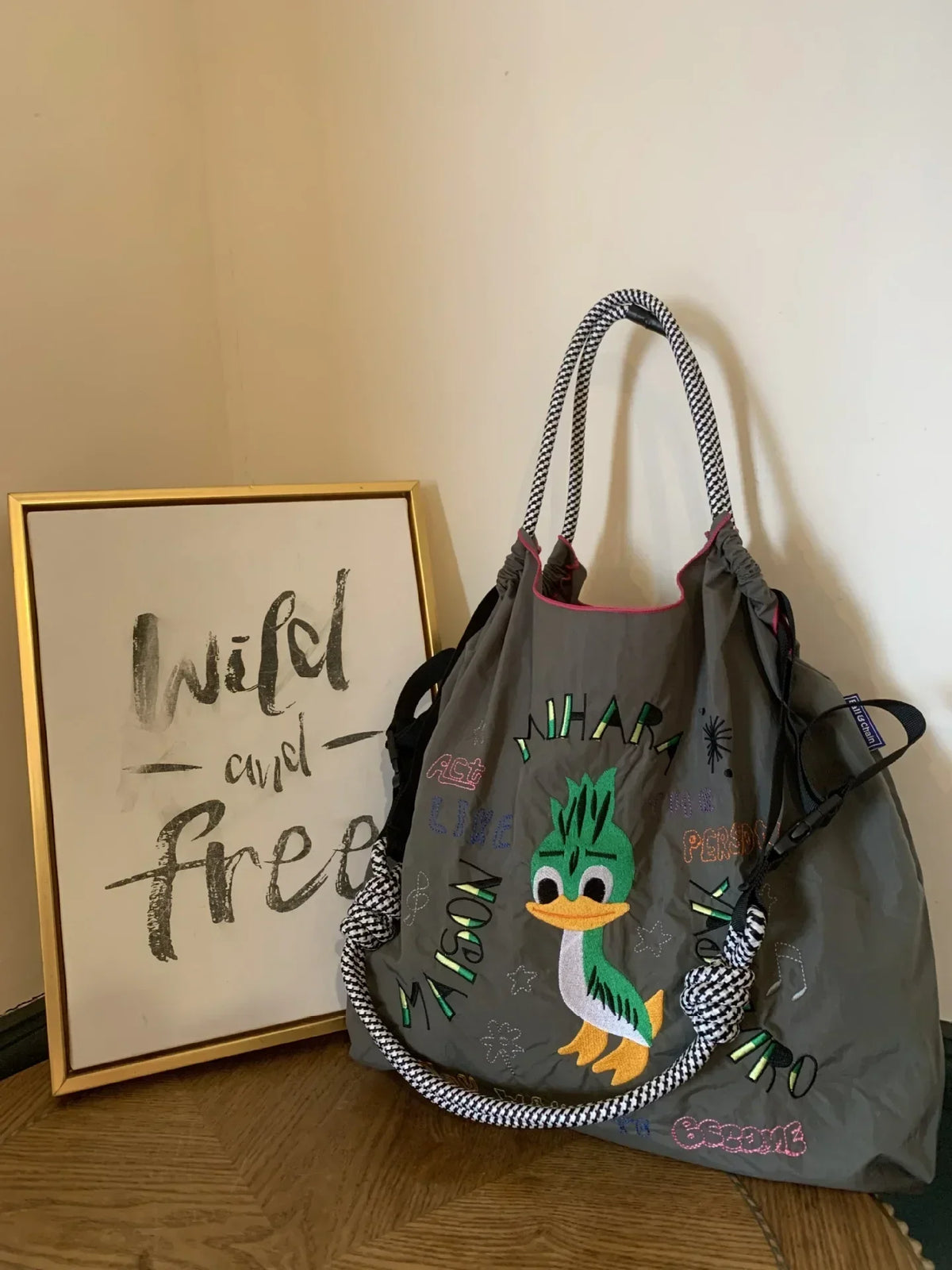 Large grey nylon tote bag with colorful embroidery featuring a duck design and playful phrases; black and white braided handles.