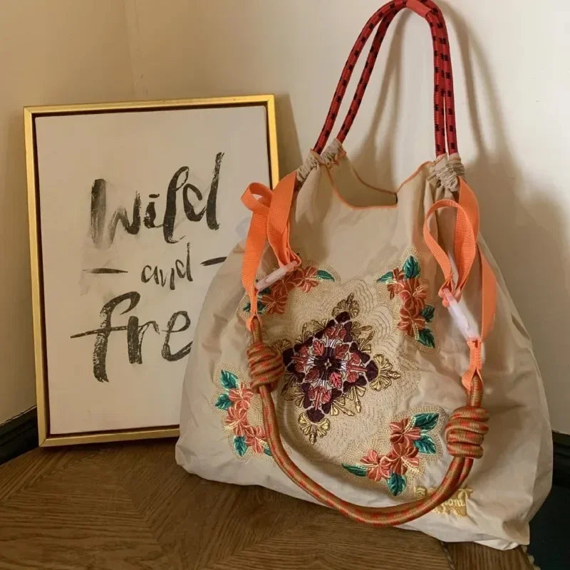 Large-capacity beige tote bag with colorful floral embroidery and orange straps, presented beside framed wall art.