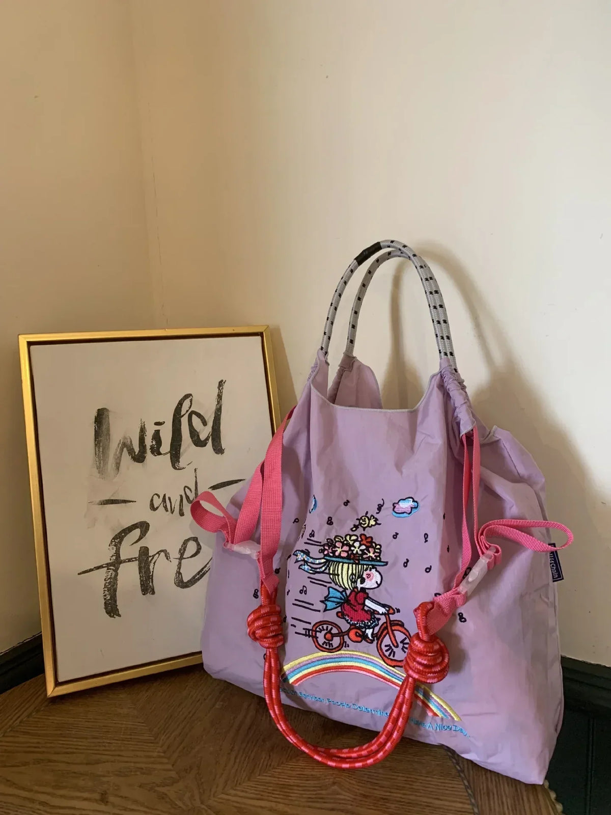 Light purple tote bag with embroidered design featuring a girl on a bicycle, pink drawstring handles, and decorated with a rainbow.