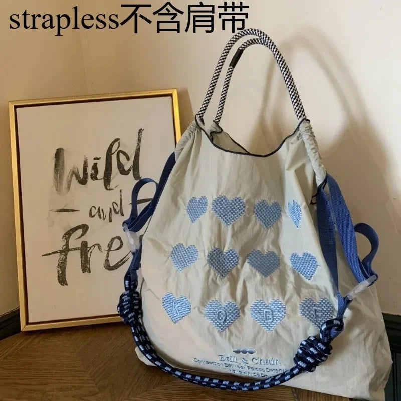 Large beige tote bag featuring blue heart embroidery, dual blue straps, and a casual design. Suitable for shopping.