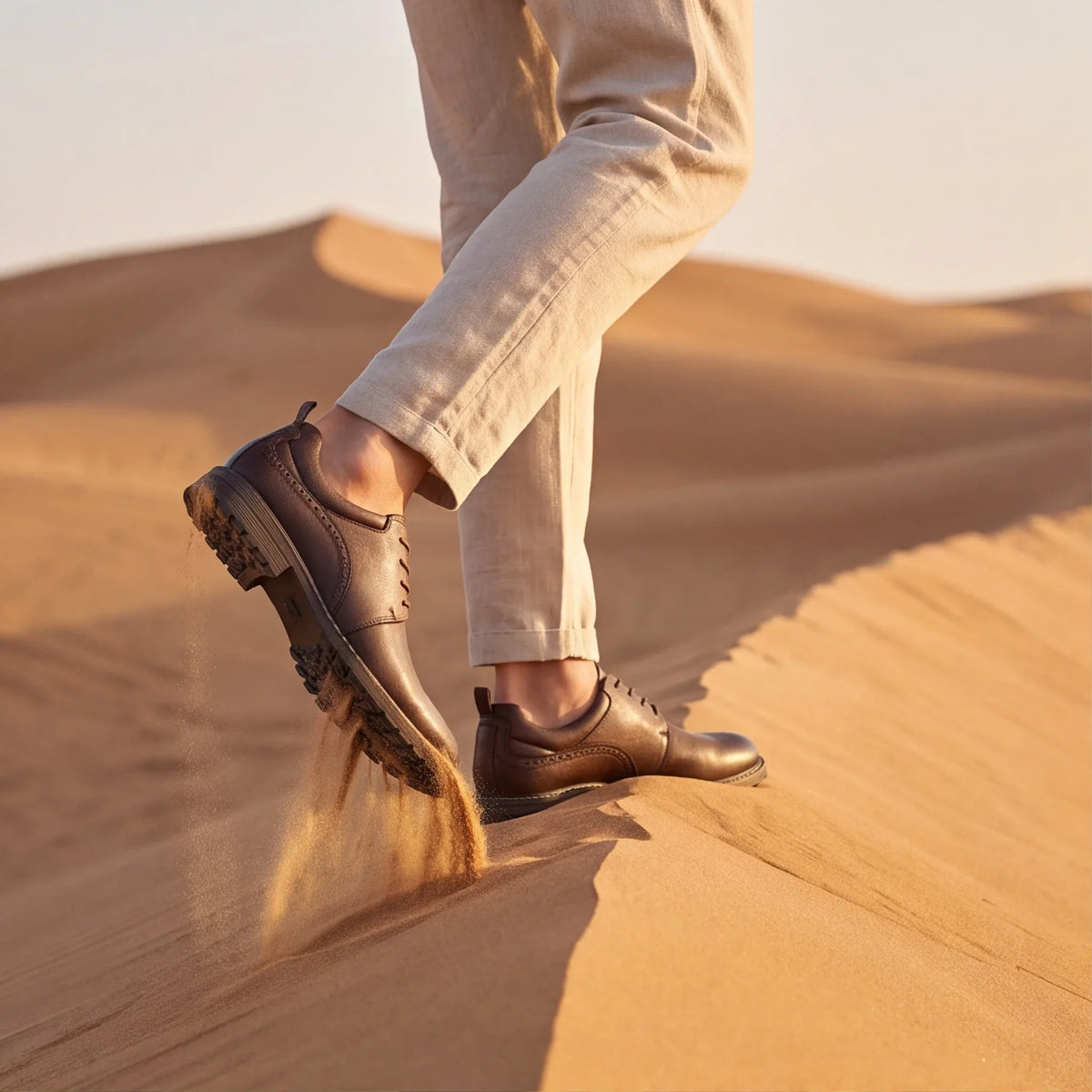 Brown lace-up derby shoes with rubber soles, worn by a model in light trousers, on sandy, uneven terrain.