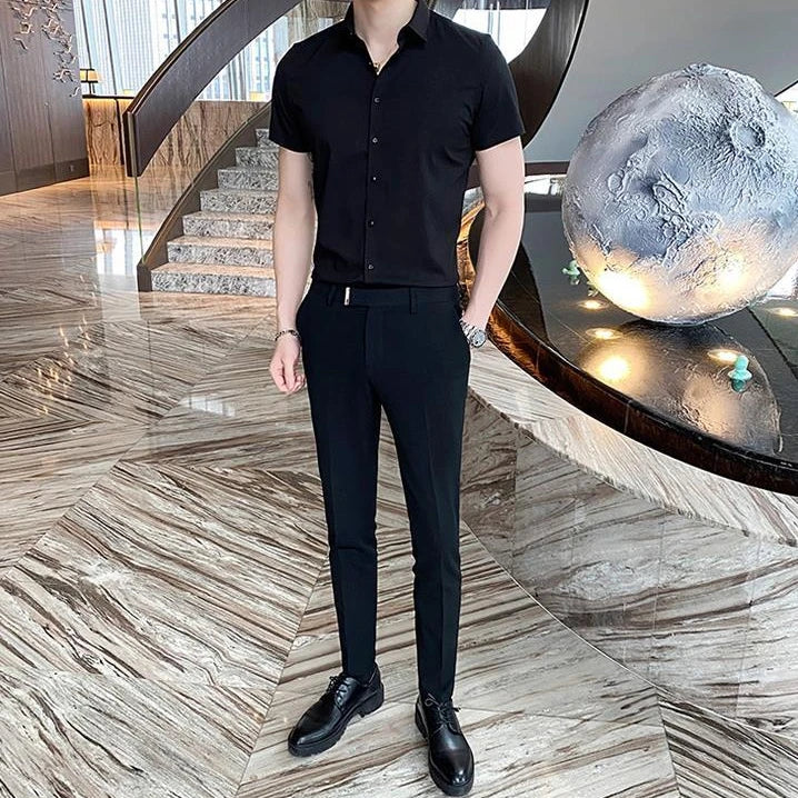 Short-sleeve black shirt with a square collar, worn tucked into slim-fit black pants. The model is standing indoors.