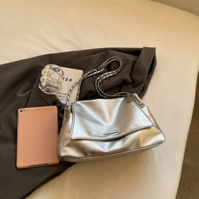 Silver shoulder bag with a soft leather finish, featuring a flap pocket and two chain straps, placed beside a tablet and book.
